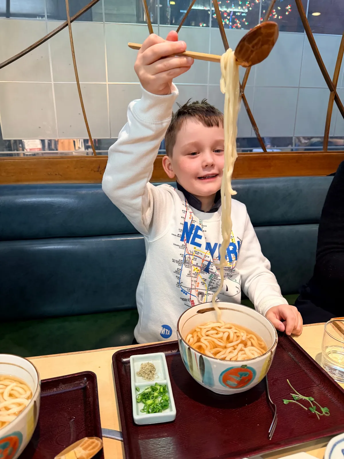 A smiling child lifts a big tangle of udon noodles with chopsticks at a Japanese restaurant.