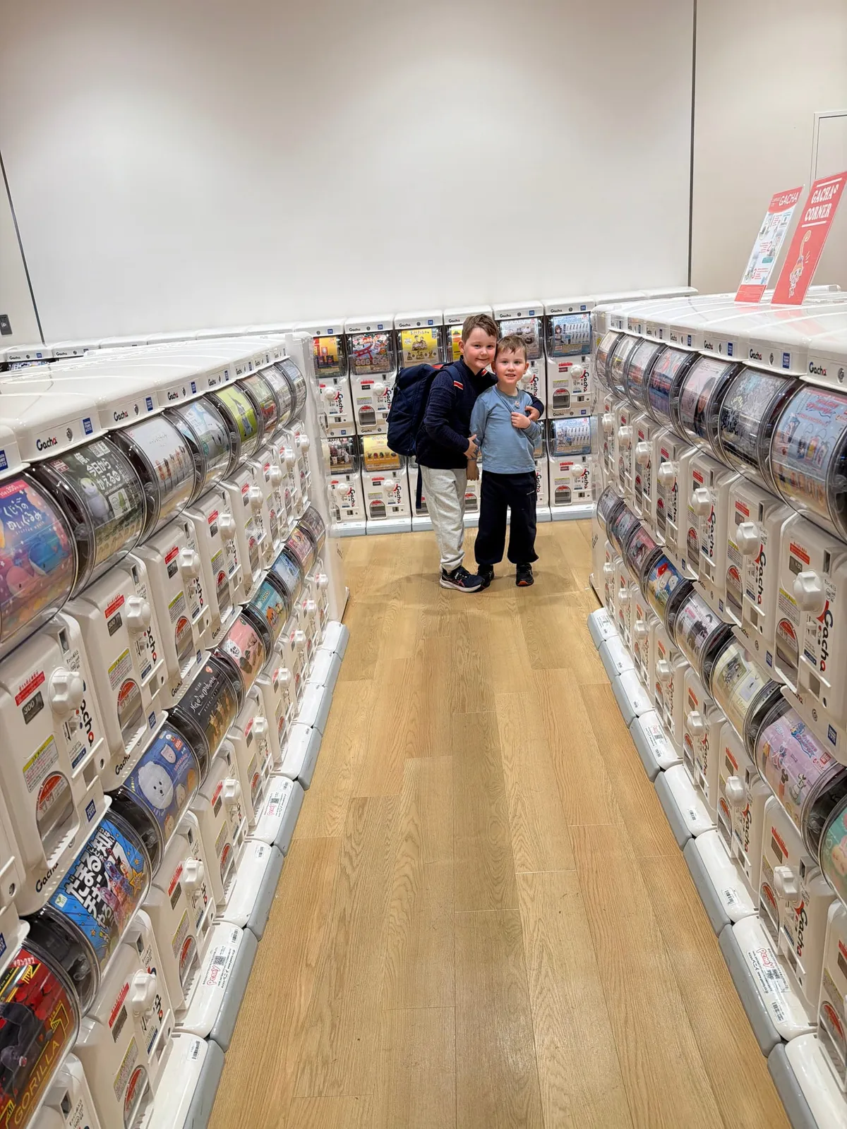 Two children hug in an aisle lined with rows of Japanese gachapon capsule-toy machines.