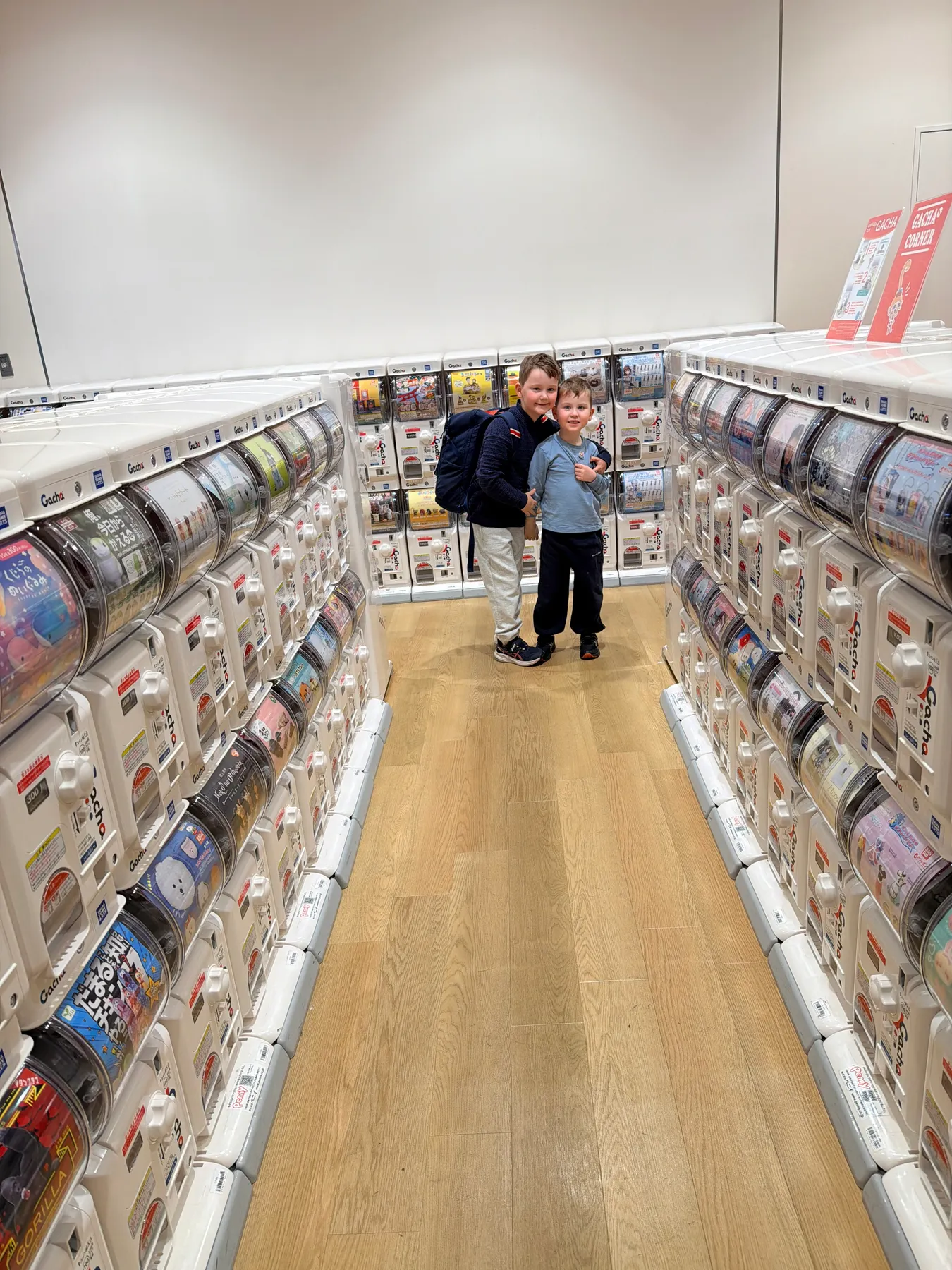 Two brothers hug while standing in an aisle lined with rows of Japanese gachapon capsule-toy machines.