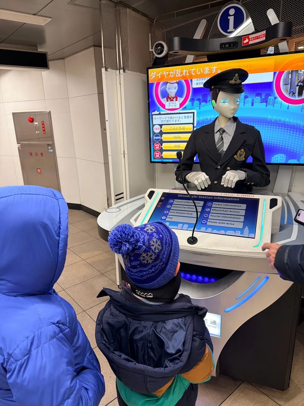 A child in a snowflake beanie looks up at a humanoid station-attendant robot at a Tokyo train station information desk.