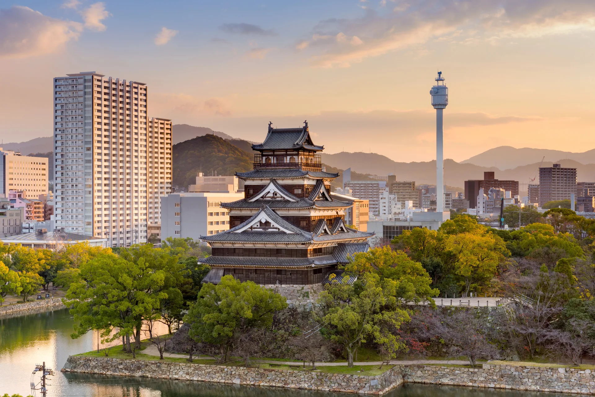 The reconstructed keep of Hiroshima Castle rising above its moat and trees at sunset.