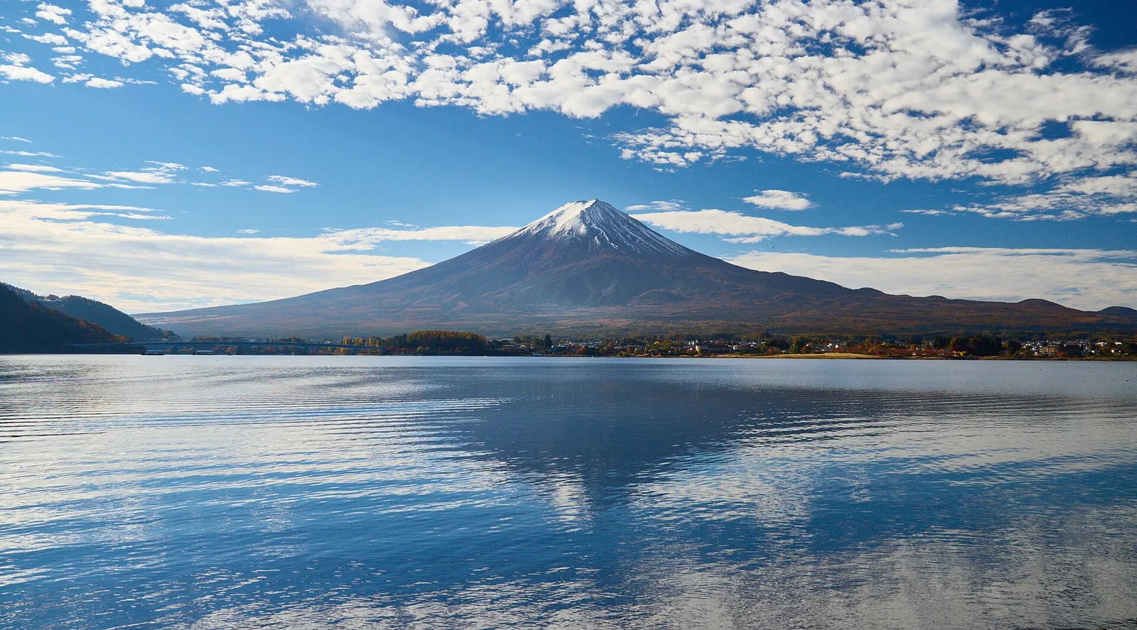 Snow-capped Mount Fuji rising above the calm water of Lake Kawaguchiko under a blue sky.