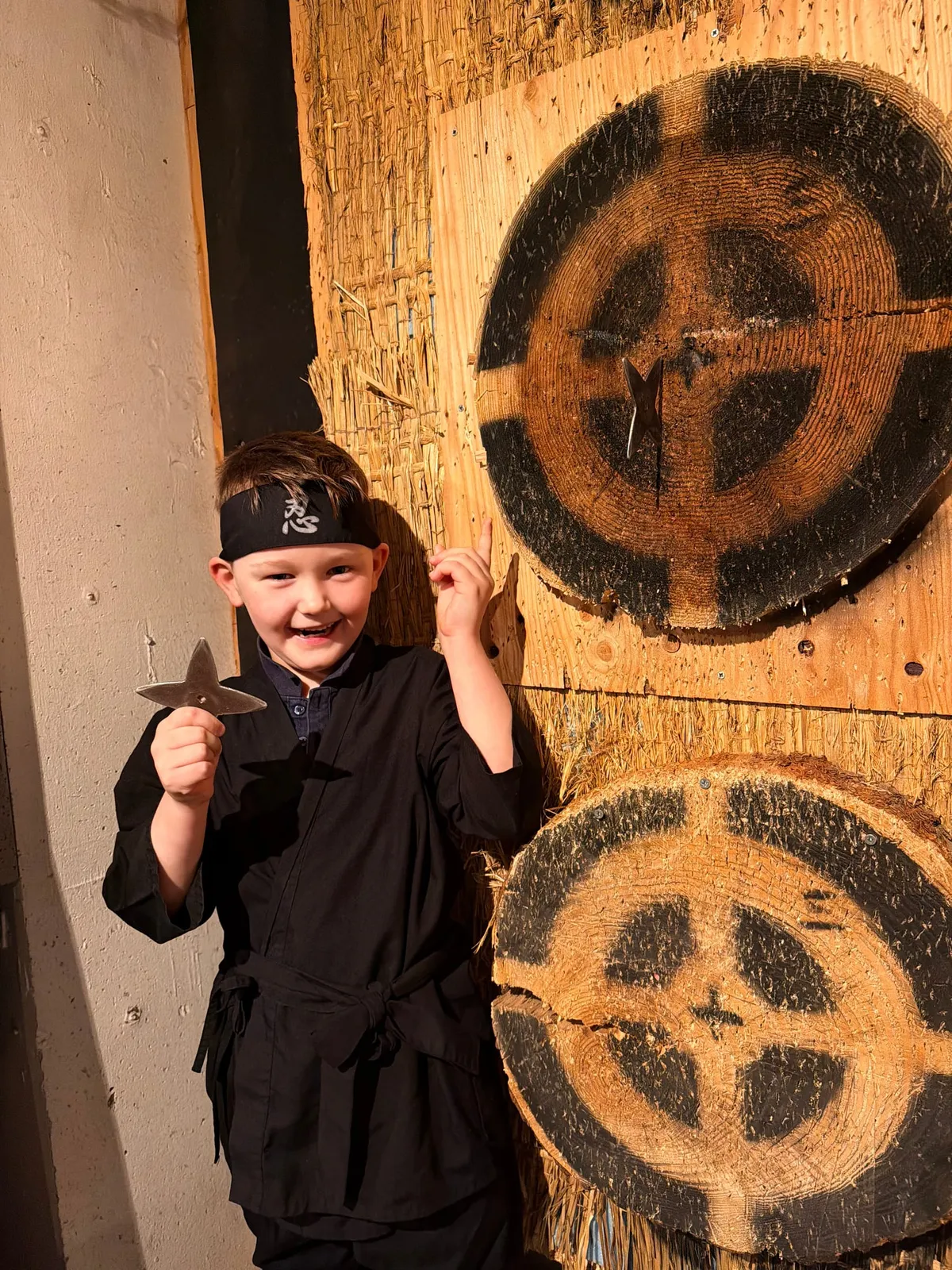 A child dressed as a ninja holds a metal throwing star in front of wooden targets at a ninja experience.