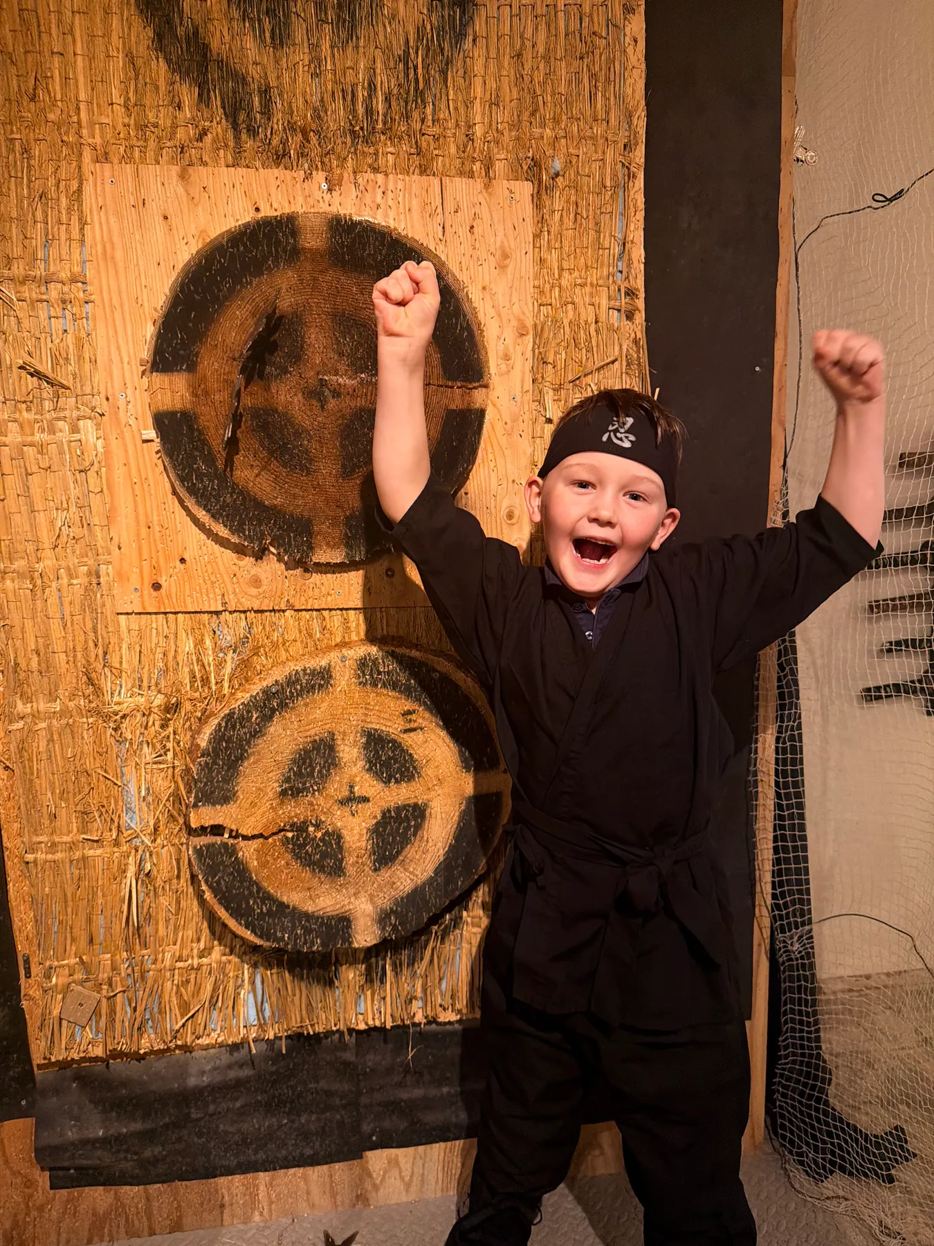 A grinning child in a black ninja costume and headband punches the air in front of wooden shuriken targets at a ninja experience.