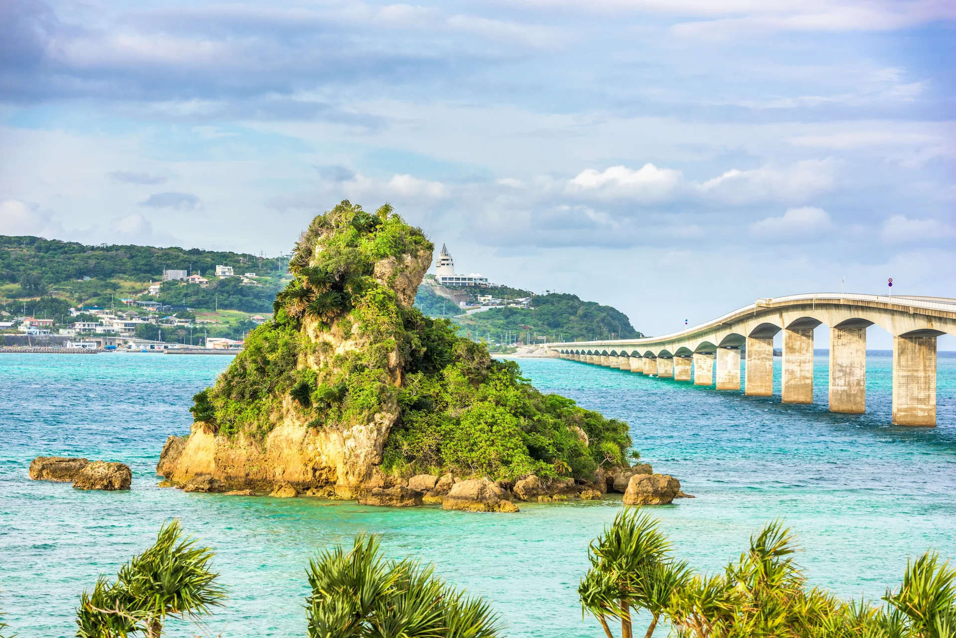 A turquoise Okinawa bay with a small green islet and a long sea bridge stretching across the water under a bright sky.