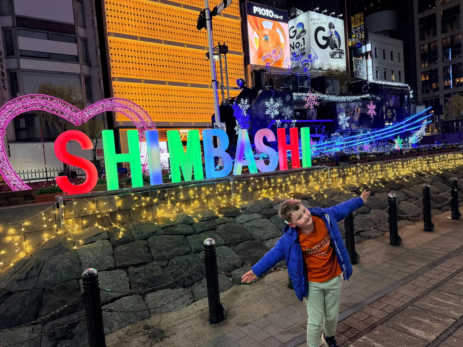 A child beside a glowing sign and an illuminated steam locomotive at Shimbashi, Tokyo, at night.