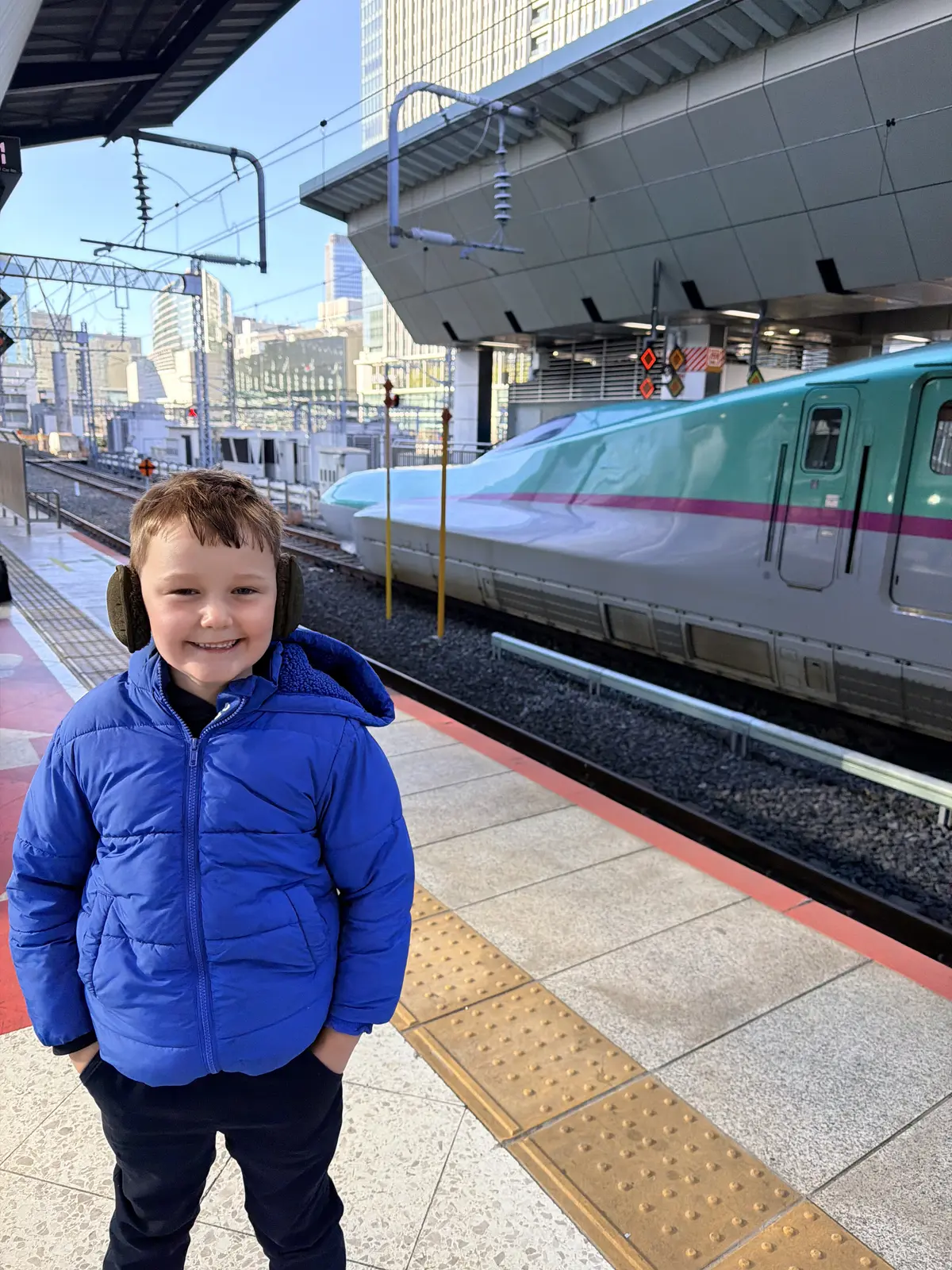 A smiling child on the platform beside a green E5-series Hayabusa shinkansen (bullet train) in Japan.