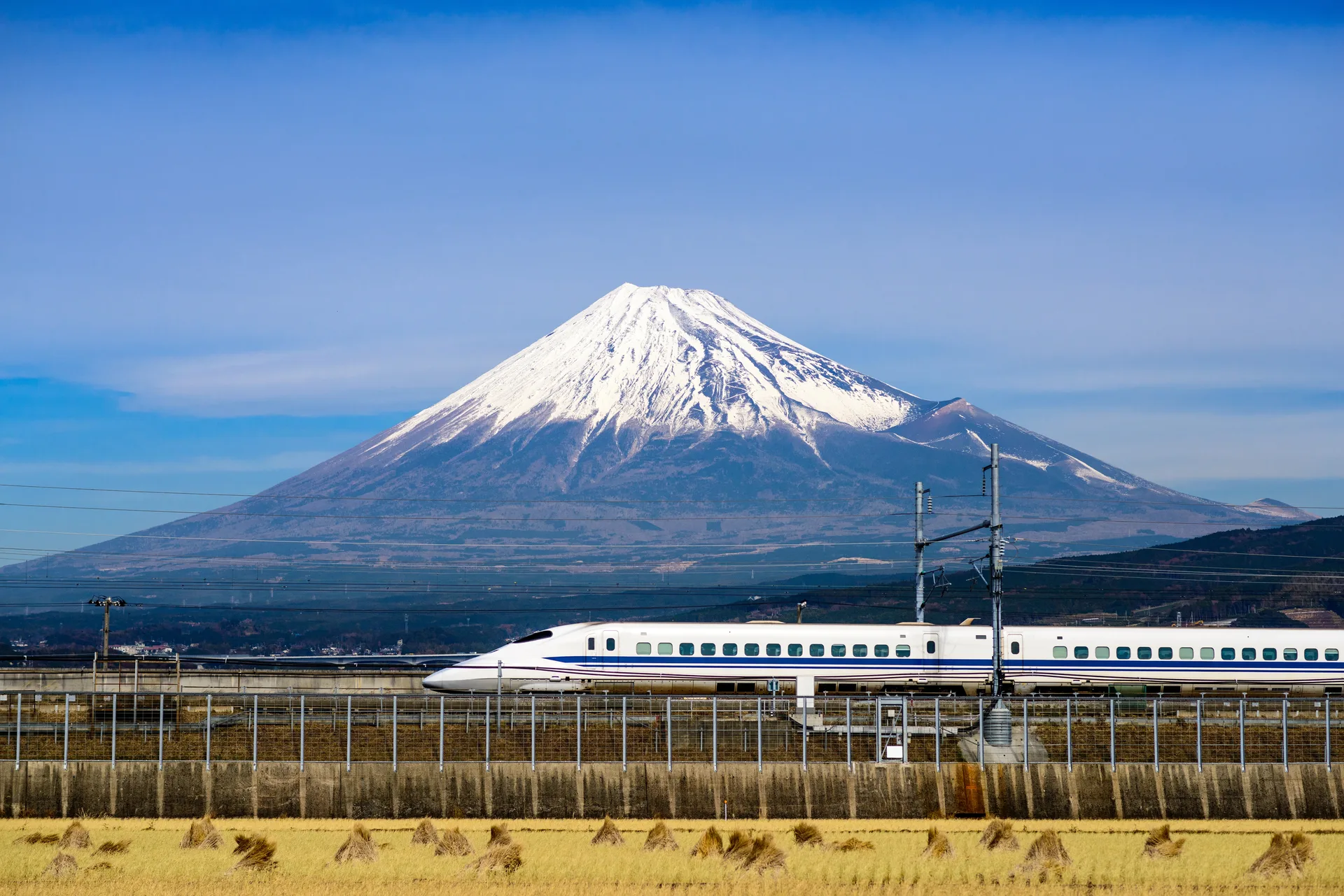 A white shinkansen bullet train passing along an embankment below the snow-capped cone of Mount Fuji.