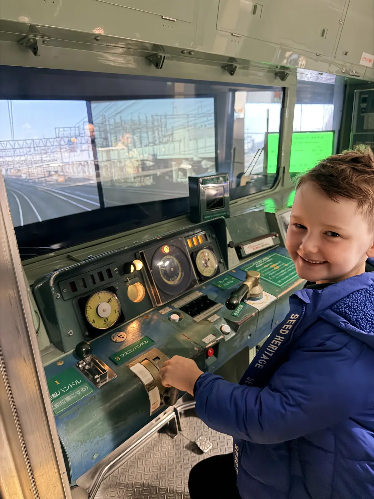 A grinning child at the front of a train watching the track ahead through the driver-cab window.